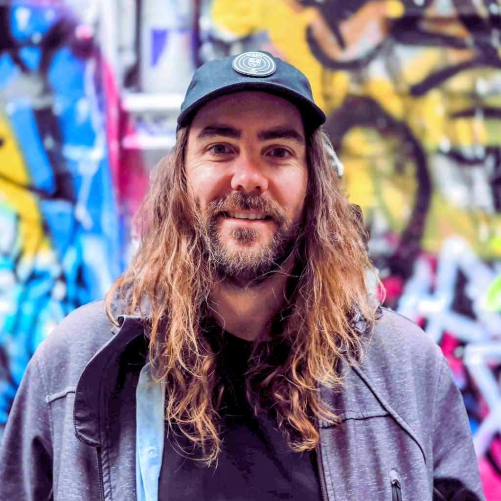 A man smiling with long brown hair, a beard and wearing a hat.